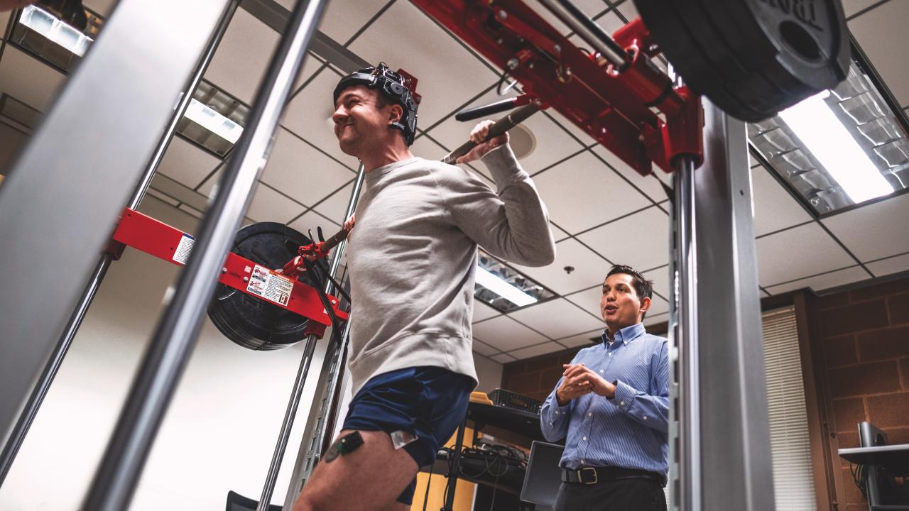 Person performing a barbell squat while wearing monitoring sensors, with a researcher observing in a lab setting.